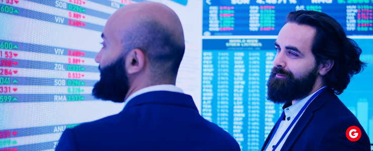 Two men in suits analyzing stock market data on a digital display in a corporate setting.